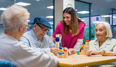 Caregiver in pink scrubs assists elderly individuals with a block-stacking activity in a brightly lit room.