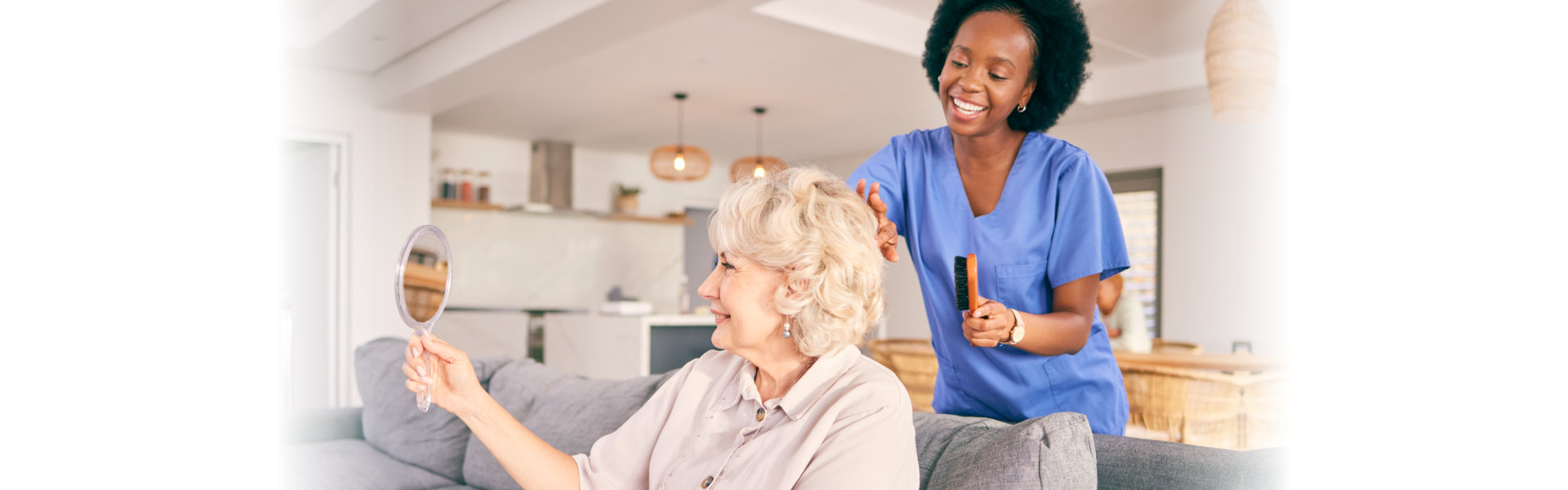 Caregiver in blue scrubs combing elderly woman's hair while she holds a hand mirror, sitting on a gray couch.